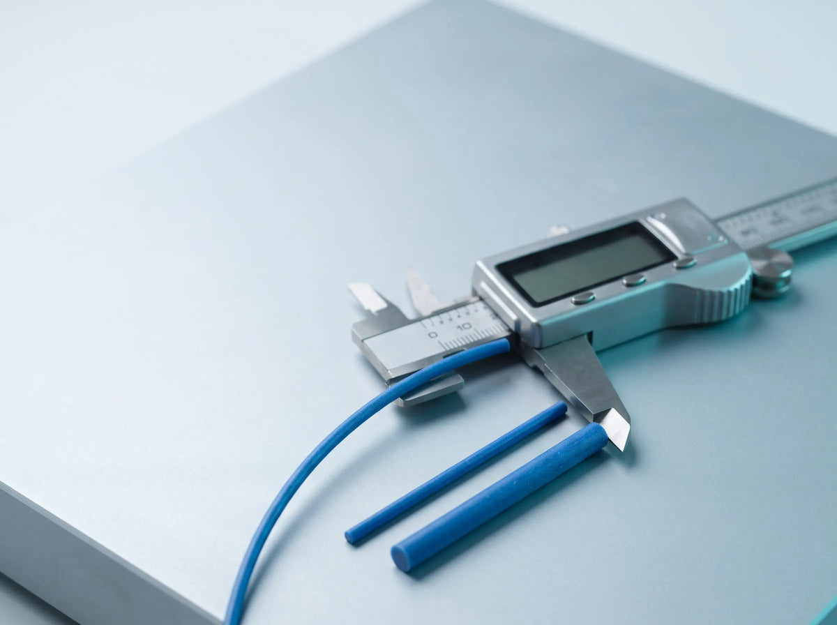 A digital caliper gently measures blue 3D printing filament on a cool white surface, with 1.75 mm and 2.85 mm filament samples shown for comparison.